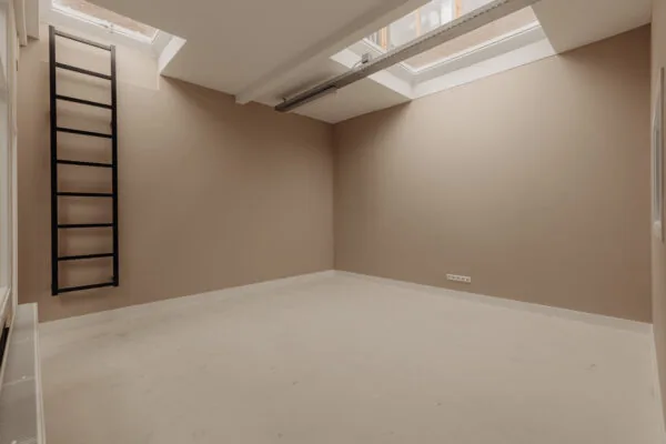 Lege kamer aan de Henri Polaklaan met beige muren, dakramen en een zwarte ladder tegen de muur.